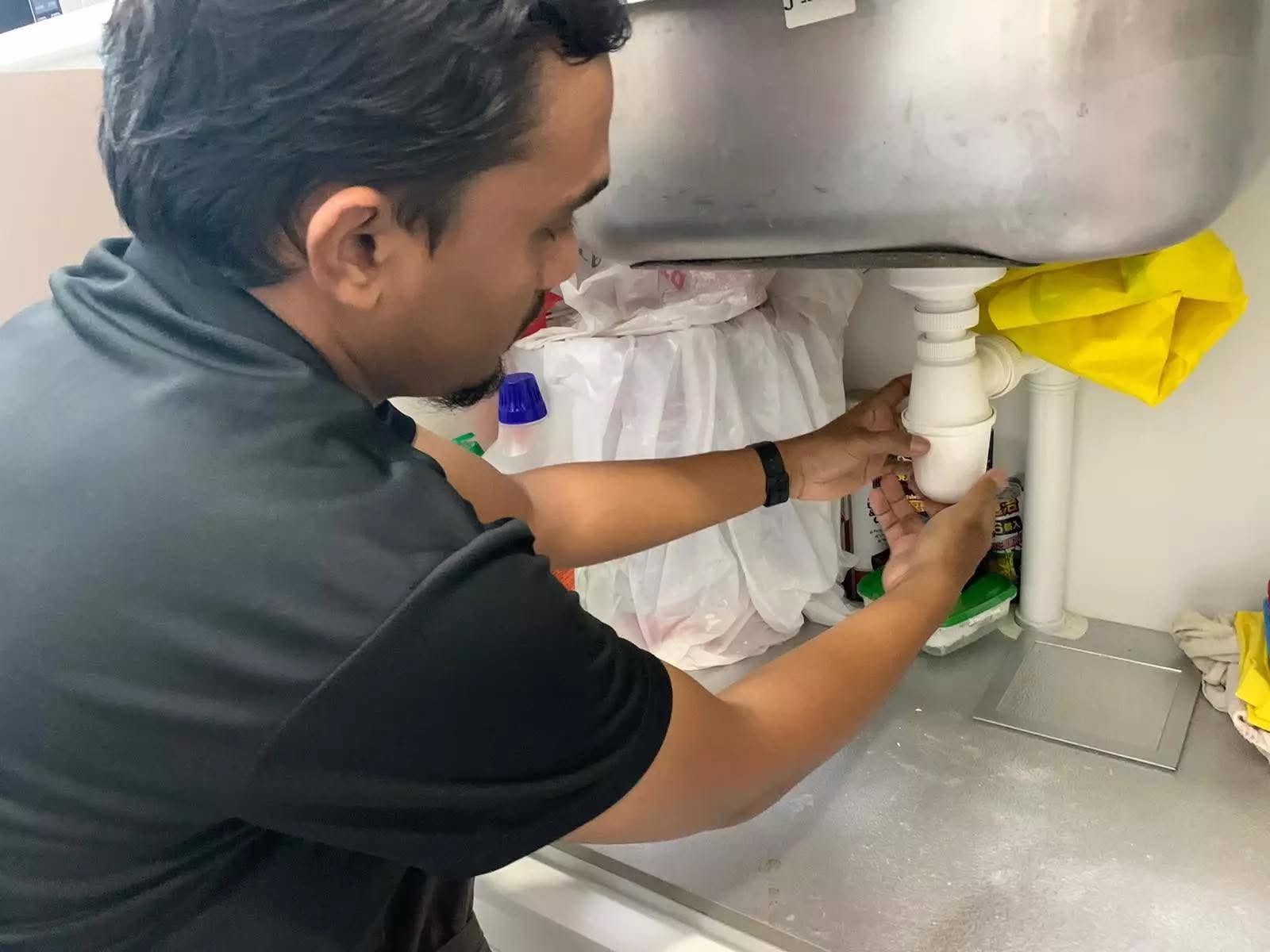 Plumber adjusting and securing the bottle trap drainage pipe underneath a stainless steel sink in Singapore.
