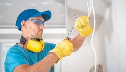 Licensed electrician wearing safety gear cutting and stripping ceiling wires for electrical repair in Singapore.