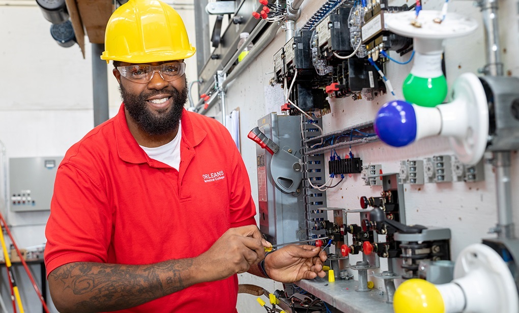 Electrician wearing a yellow hard hat and red shirt working on a complex electrical circuit test board with light fixtures and switches.