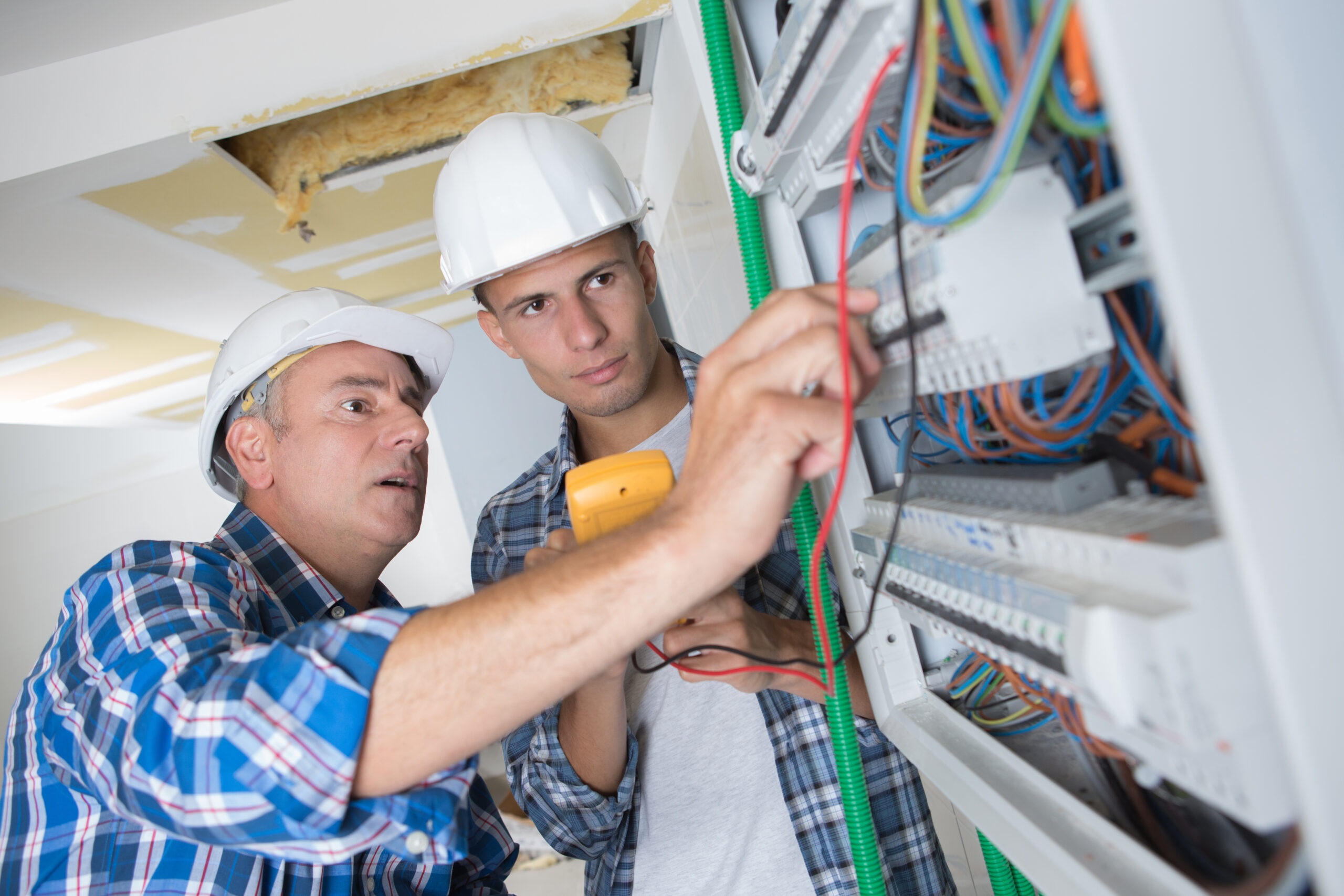Two electricians wearing hard hats inspecting and testing circuit breaker wiring in an open electrical distribution board.