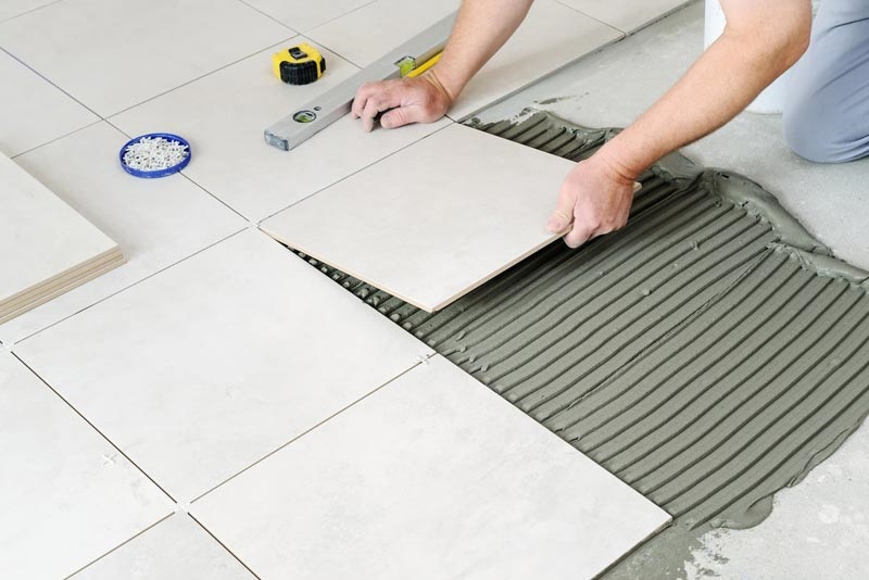 Contractor hands laying off-white ceramic floor tiles onto notched adhesive mortar bed with measuring tools nearby.