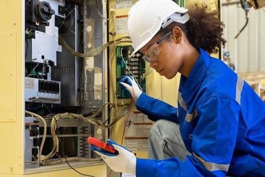 Female technician wearing a white hard hat, safety glasses, and blue workwear using a digital multimeter to inspect an industrial machinery control panel.