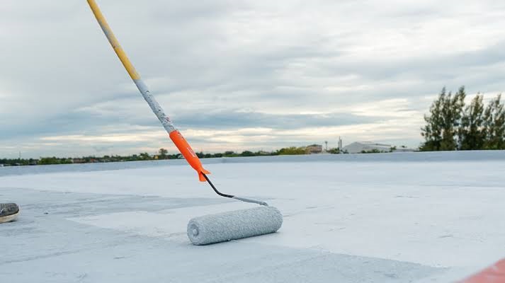 Long-handled paint roller applying white liquid elastomeric waterproofing membrane coating on a flat rooftop floor.