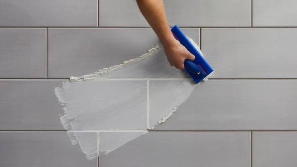 Hand using a rubber grout float to spread white tile grout into joints across light grey rectangular floor tiles.