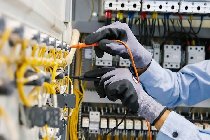 Electrician wearing protective work gloves using multimeter probes to test voltage on terminal blocks within an electrical panel.