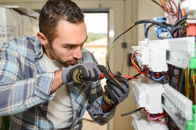 Electrician wearing work gloves using wire strippers on cables connected to an electrical circuit breaker panel.