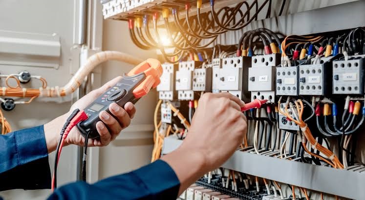 Electrician using a digital clamp meter probe to inspect voltage and wiring in an electrical distribution panel.