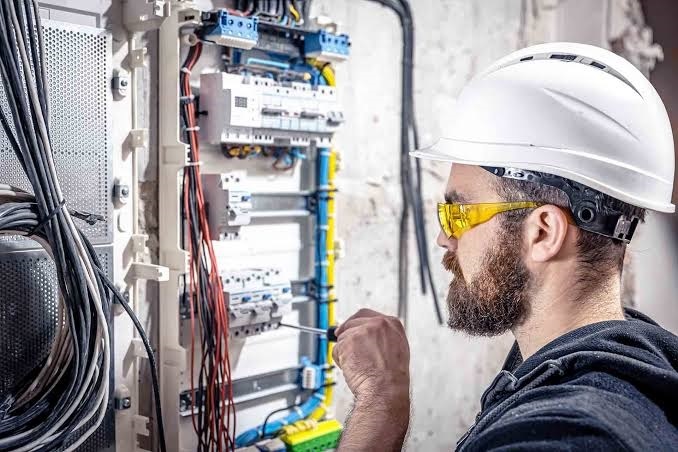 Electrician wearing a white hard hat and yellow protective safety glasses working on a main electrical breaker panel with a screwdriver.