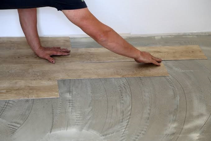 Flooring installer hand-laying wood-grain vinyl floor planks over notched adhesive substrate on subfloor.