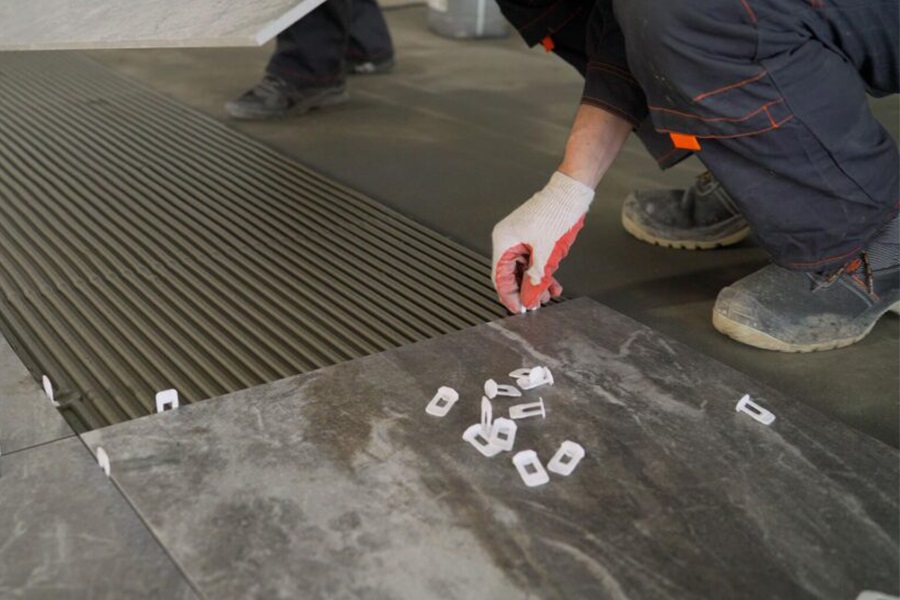 Tiler installing dark grey floor tiles using plastic leveling clips and spacers over comb-combed adhesive mortar.